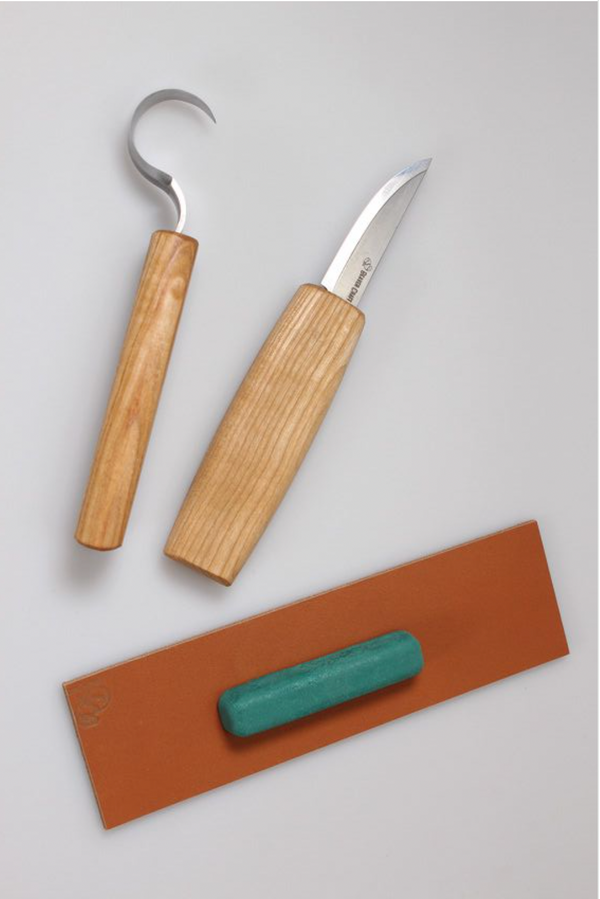 A set of wooden spoon carving tools including a small whittling knife, a spoon carving knife, a leather strop, and a green polishing compound, displayed on a white background.